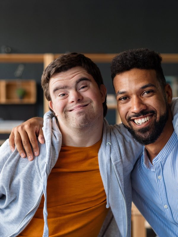 Happy young man with Down syndrome and his tutor with arms around looking at camera indoors at school