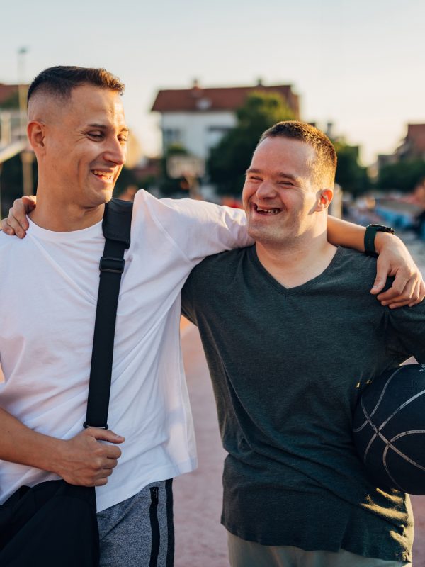 Cheerful man with Down syndrome hugs his cheerful best friend as they walk on an outdoor sports court, ready to play basketball
