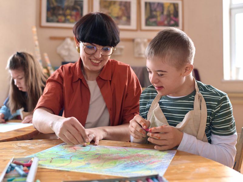 Portrait of smiling boy with disability enjoying art class in studio with mentor assisting